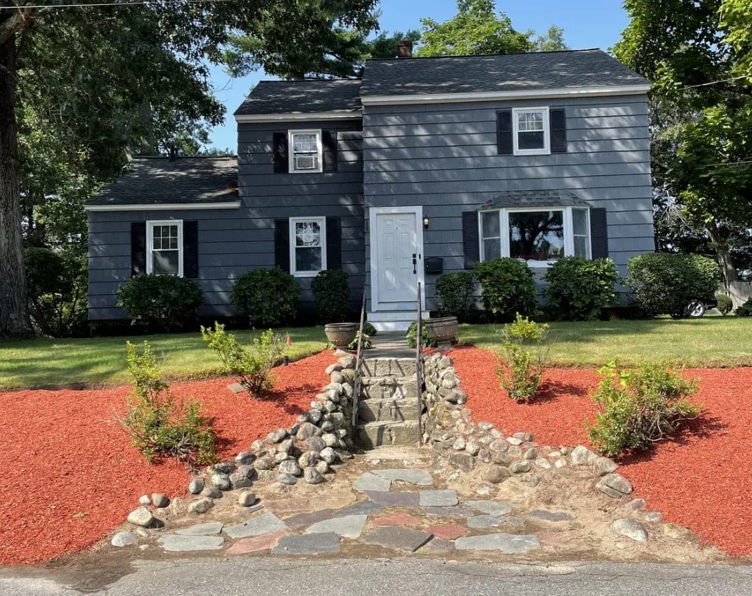 Front Yard Landscape Transformation after - An overgrown, weed-choked front yard cleared and rebuilt — fresh mulch beds, new plantings, and a clean natural-stone walkway framing the entry.